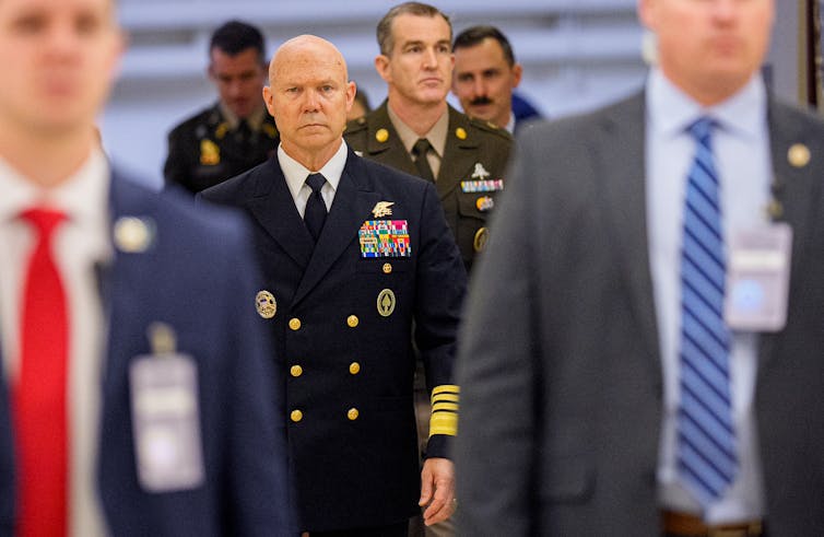 A man in a dark blue military uniform walks among a number of men in suits.