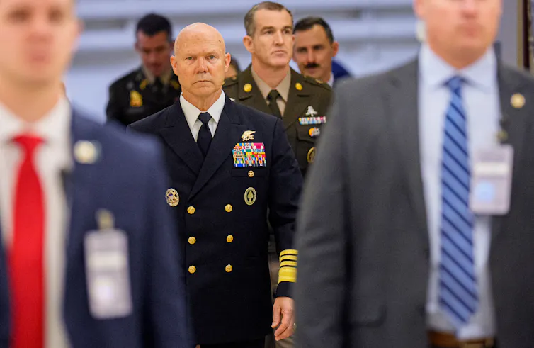 A man in a dark blue military uniform walks among a number of men in suits.