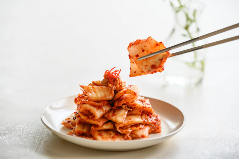 Um par de pauzinhos de metal pega kimchi de uma tigela branca.
