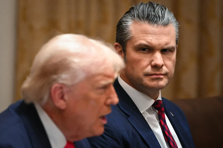 Two men in suits at a meeting, one talking, the other looking at him.