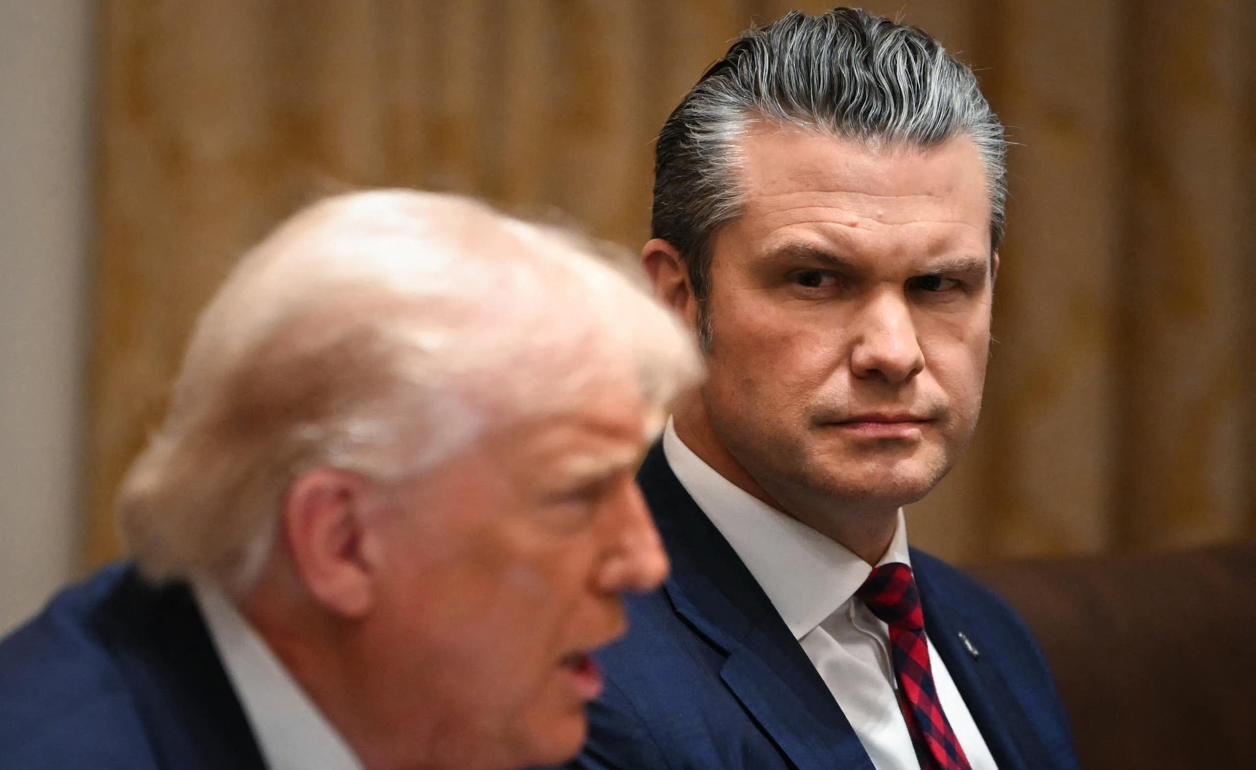 Two men in suits at a meeting, one talking, the other looking at him.