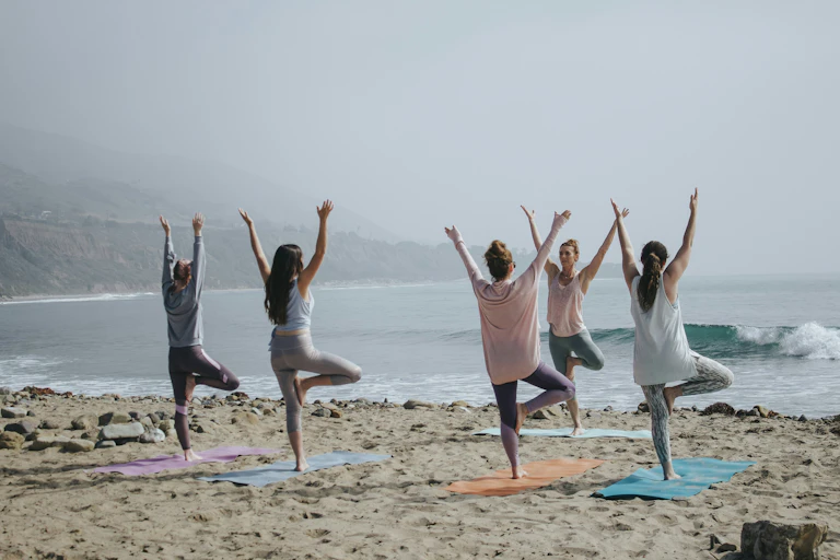 A group of women do yoga on a beach, with towels splayed out beneath them and a haze ocean backdrop.