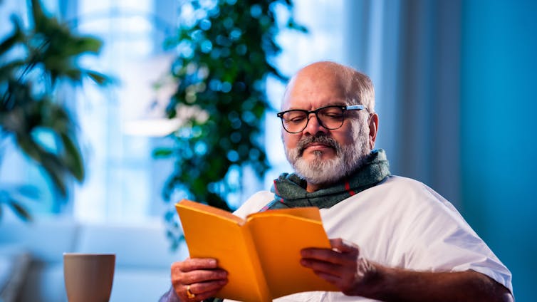 A bald, middle-aged man with a white beard reads from an orange book.