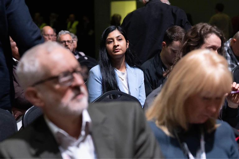 Zarah Sultana sitting several rows behind Jeremy Corbyn in an audience.