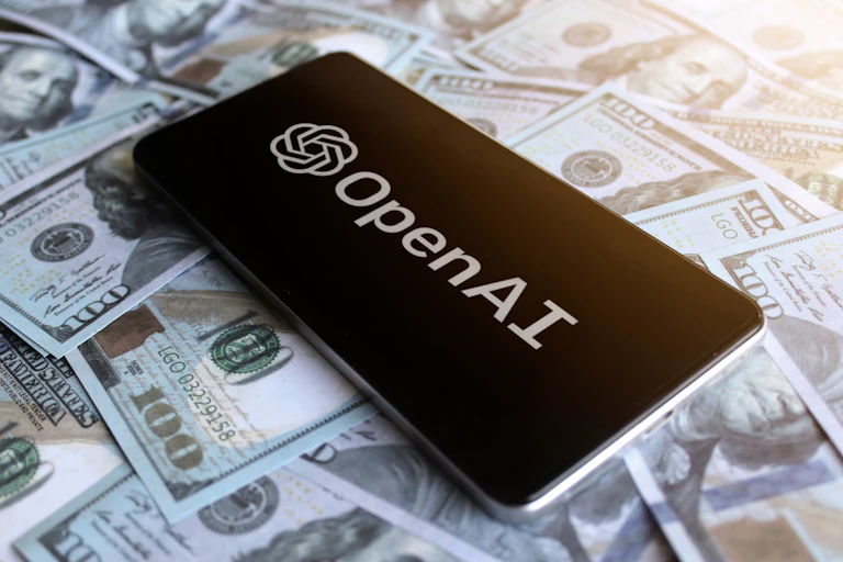 Phone showing OpenAI logo on top of US$100 bills.