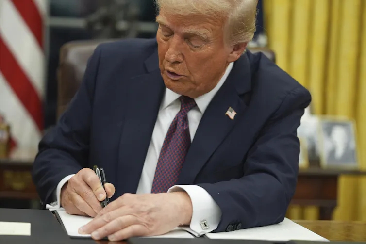 A seated man in a suit and tie signs a document.