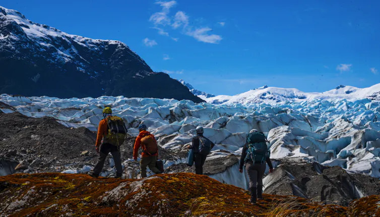 People walk along a mountain ridge with a glacier in the background.