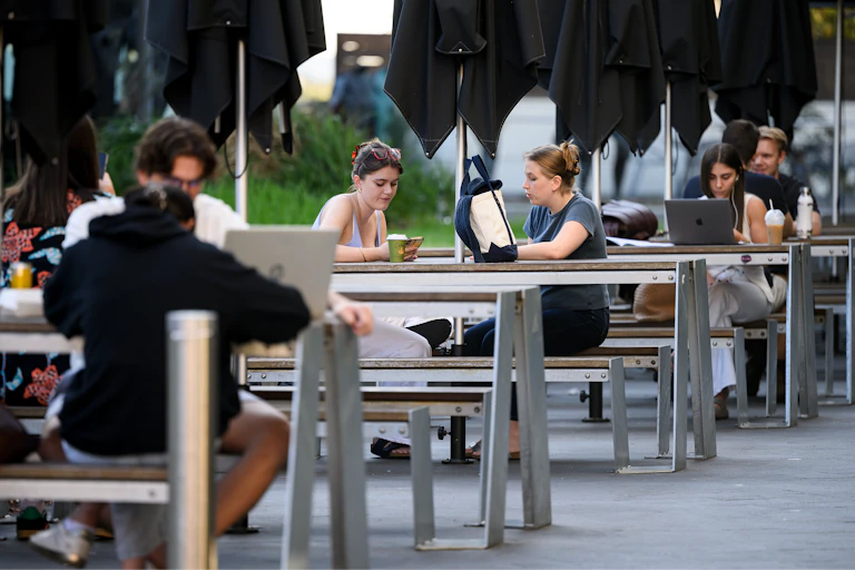 Young people sit with laptops at outdoor benches at Sydney University.