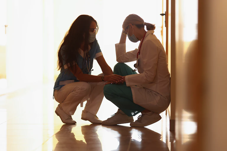 Two middle-aged health professionals kneeling in a hallway, with one comforting the other.