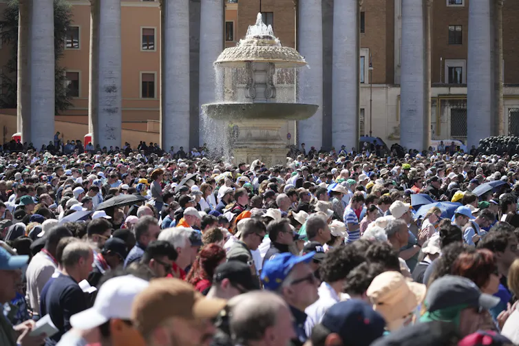A large crowd with bowed heads gathers near a fountain, beside tall white columns.