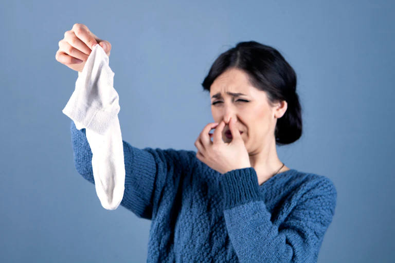 A woman plugs her nose and grimaces, looking at a smelly white sock she's holding in her other hand.