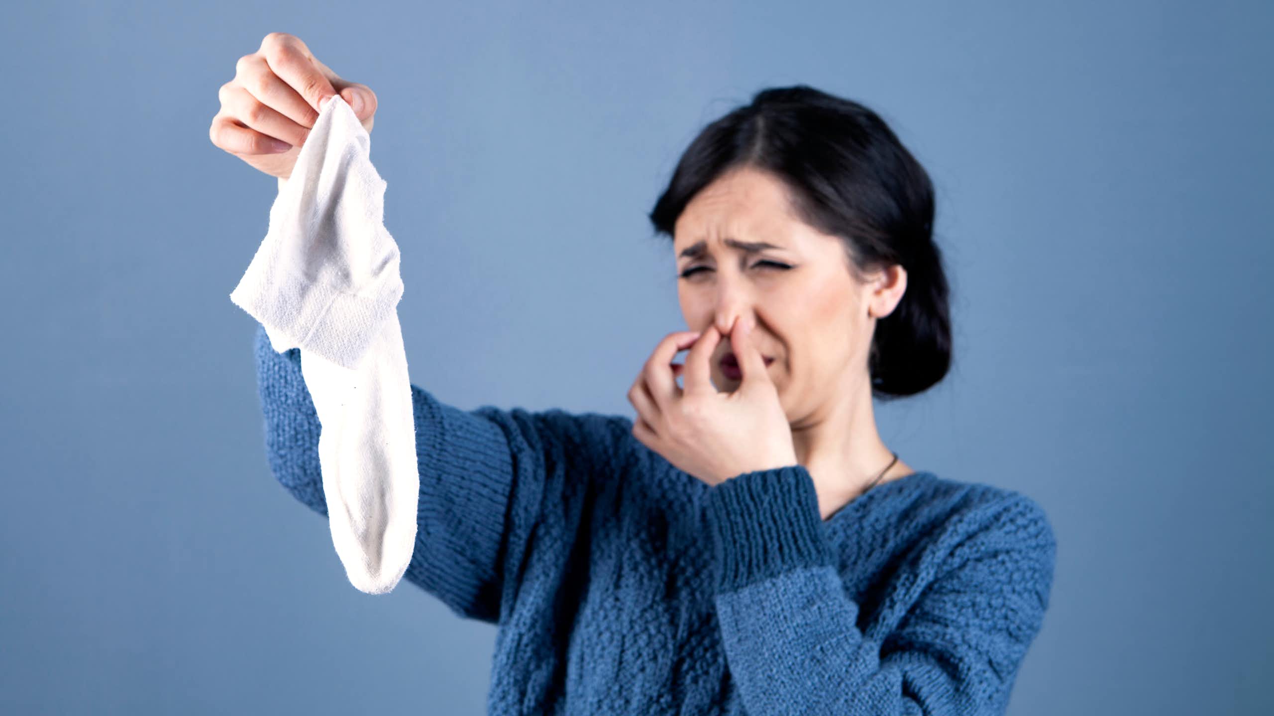 A woman plugs her nose and grimaces, looking at a smelly white sock she's holding in her other hand.