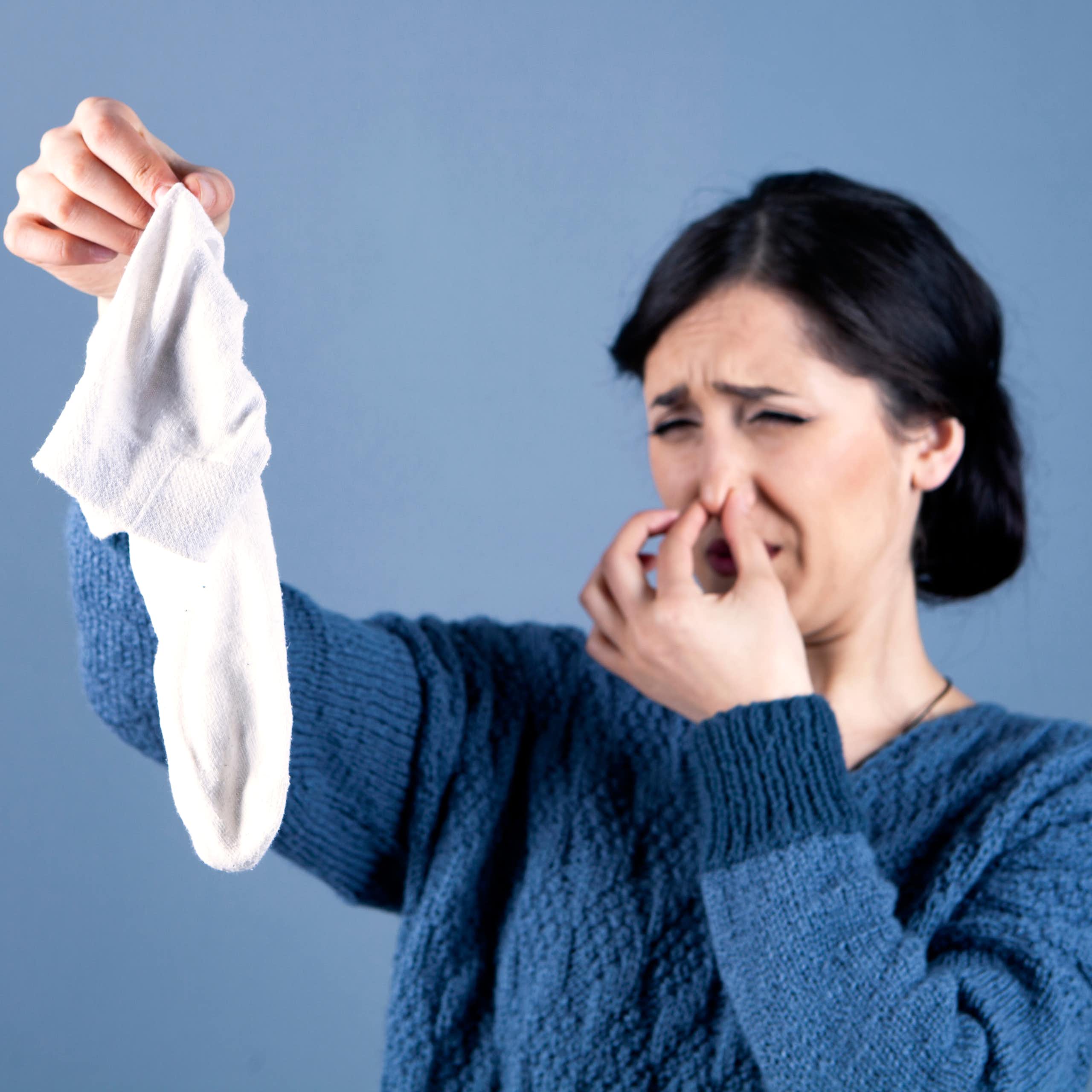 A woman plugs her nose and grimaces, looking at a smelly white sock she's holding in her other hand.