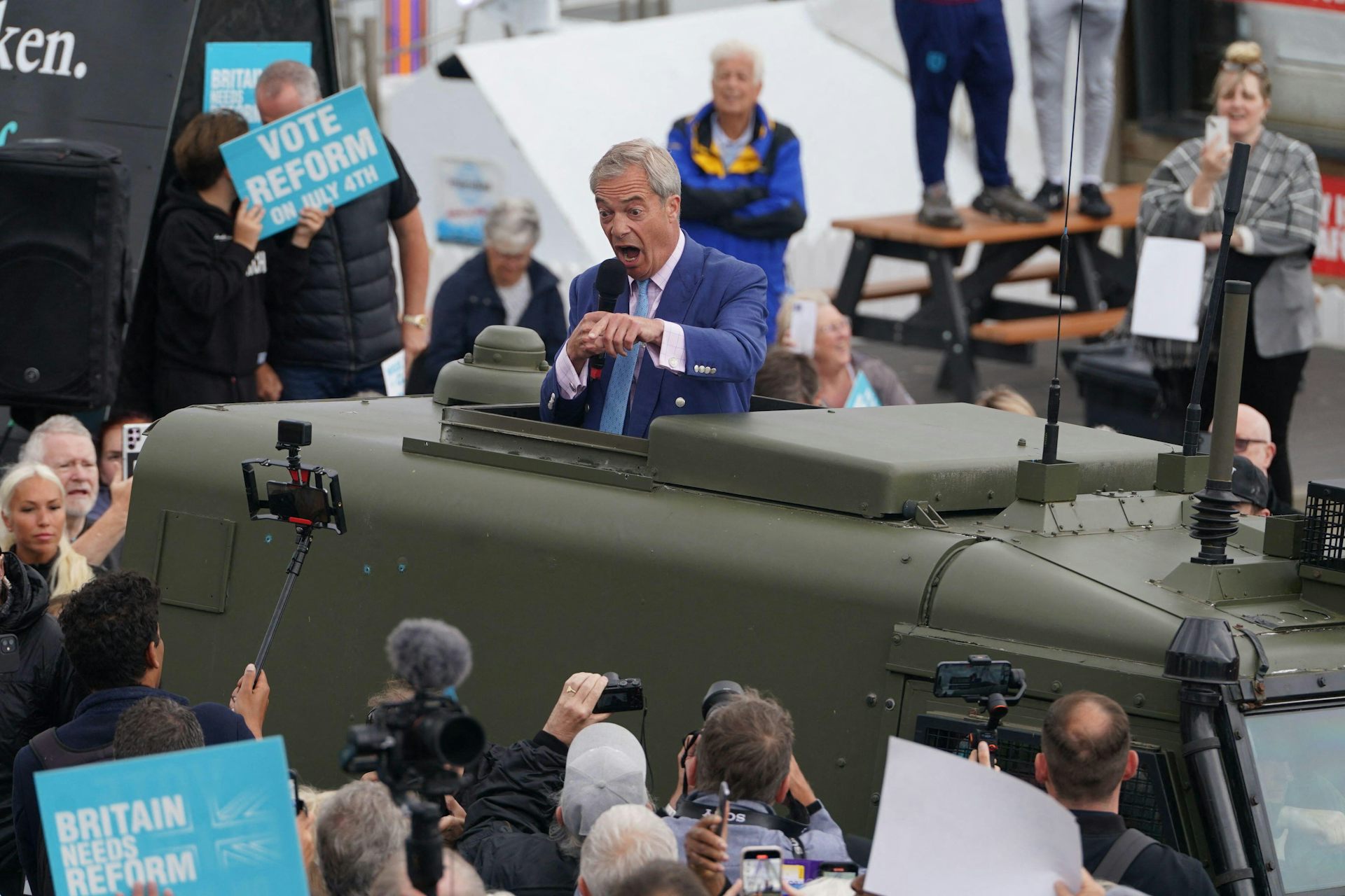Nigel Farage yelling into a microphone while standing in an open topped armoured vehicle. 