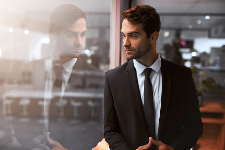 A man wearing a business suit stands next to a glass wall with his image appearing in it.