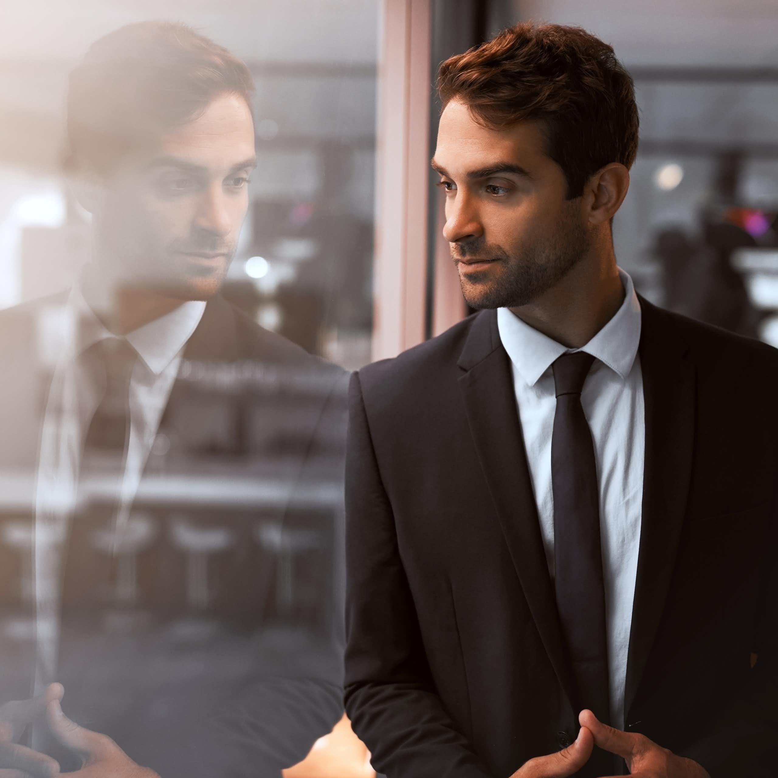 A man wearing a business suit stands next to a glass wall with his image appearing in it.
