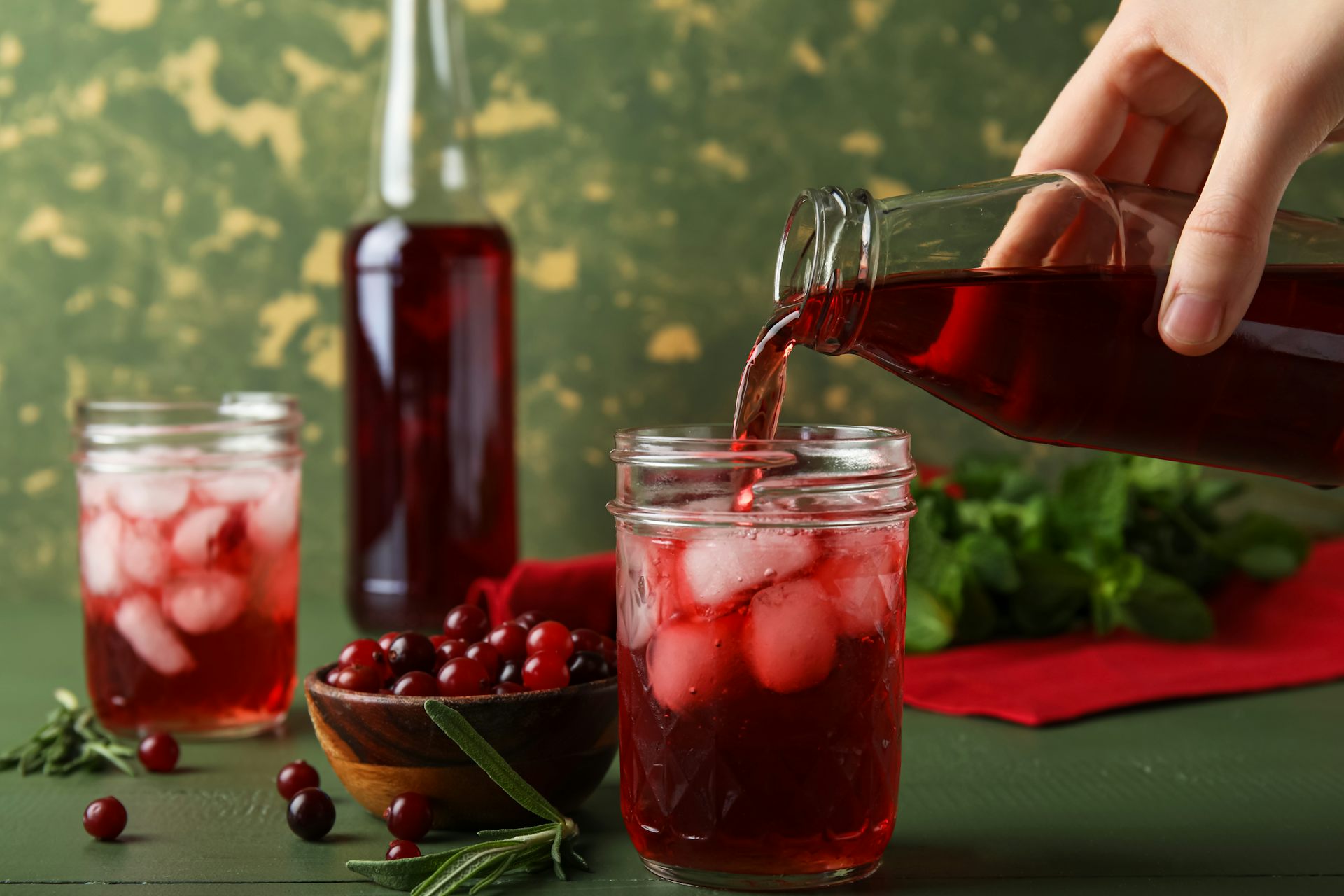 Hand pouring cranberry juice into a glass with ice cubes. A bowl of fresh cranberries is nearby.