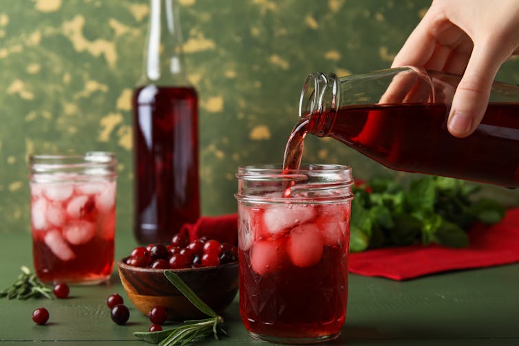 Hand pouring cranberry juice into a glass with ice cubes. A bowl of fresh cranberries is nearby.