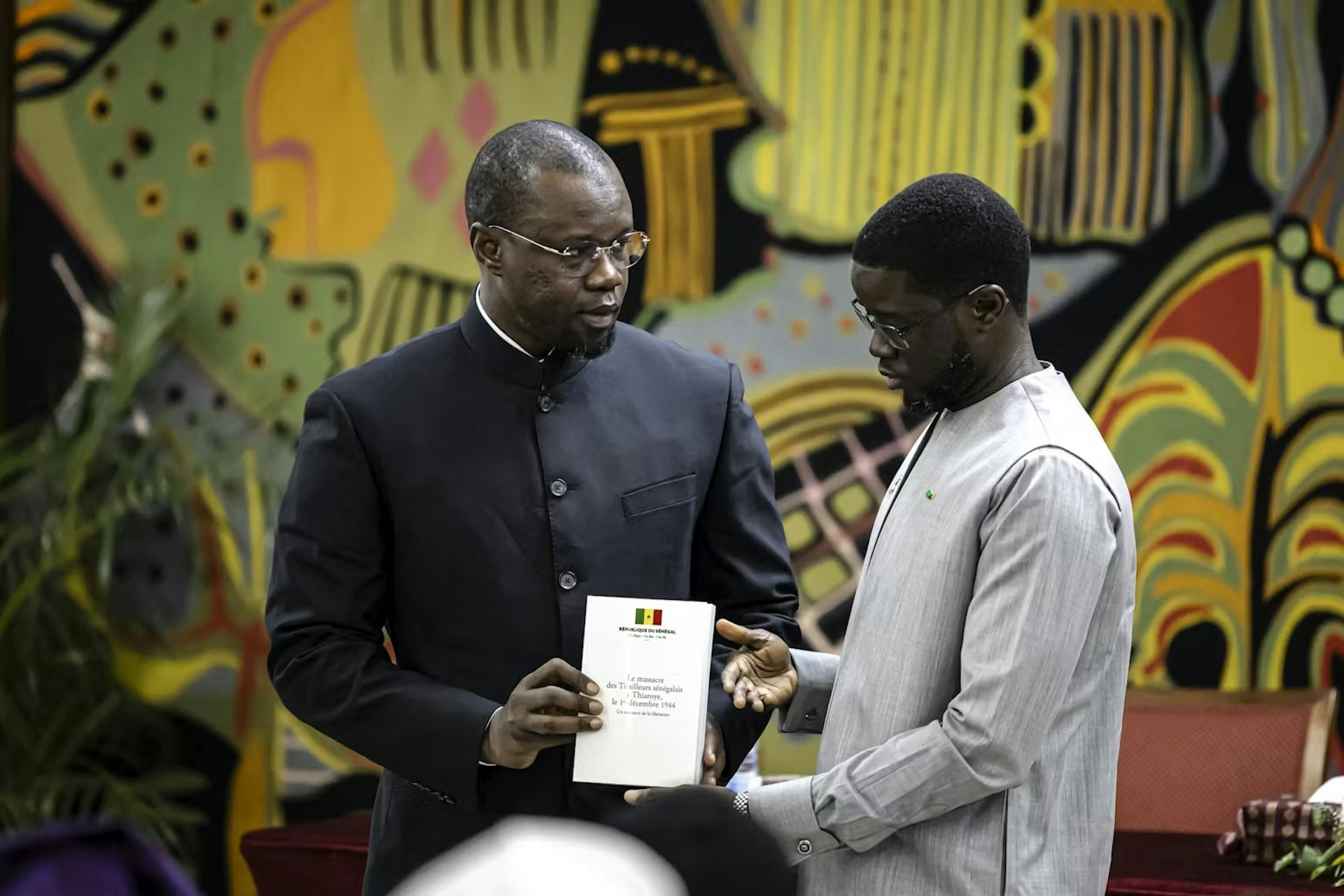 Two African men stand against a colourful art backdrop and hold a document between them.