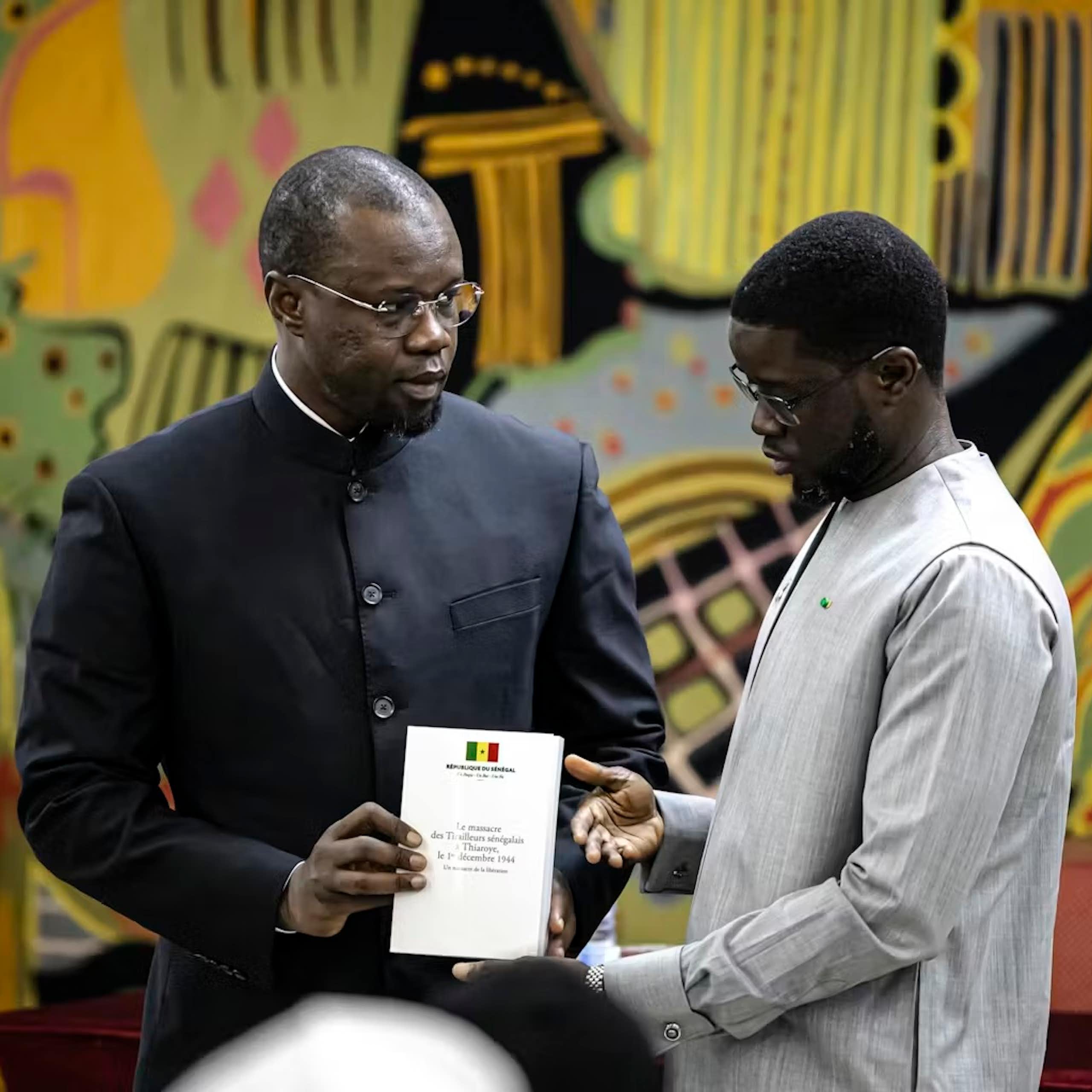 Two African men stand against a colourful art backdrop and hold a document between them.