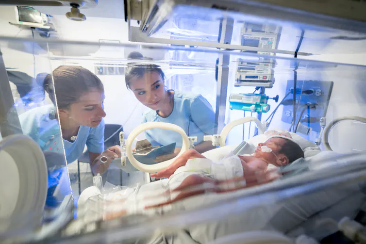 Two nurses look at an infant lying in an incubator.