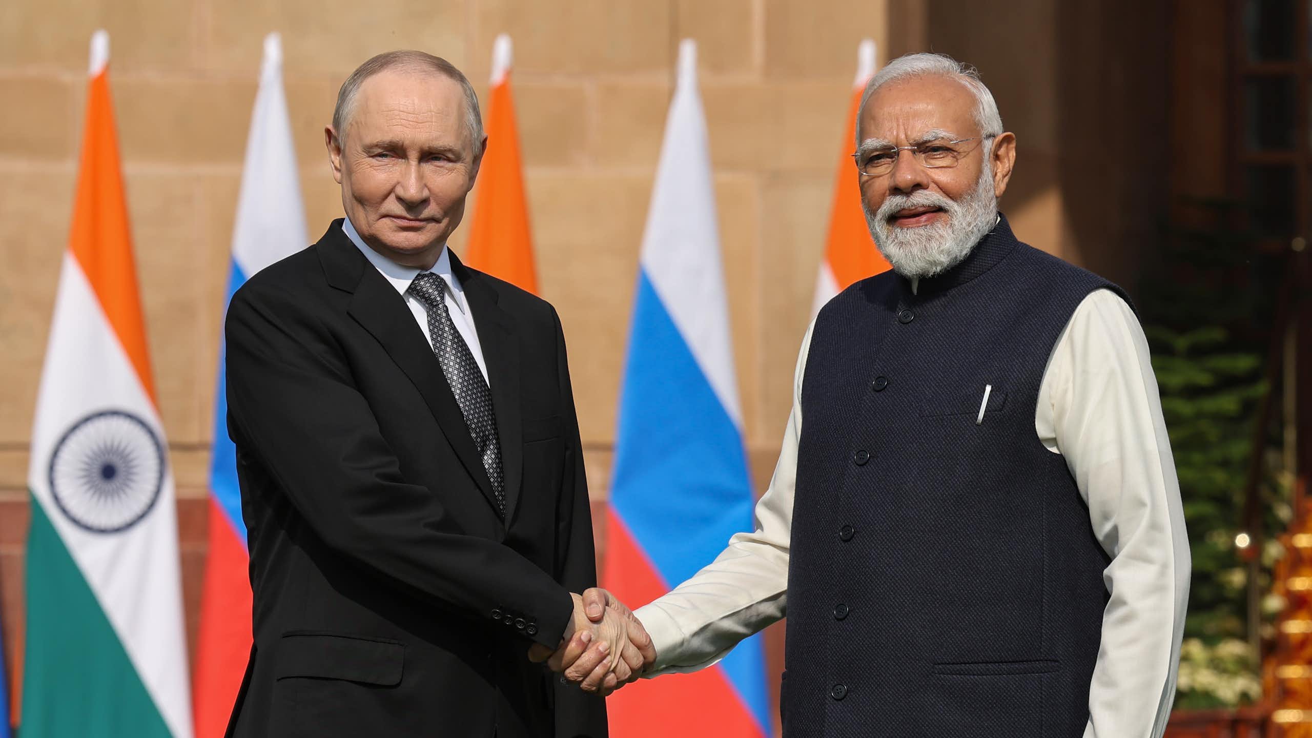 Putin and Modi, outdoors, shake hands in front of a row of Indian and Russian flags