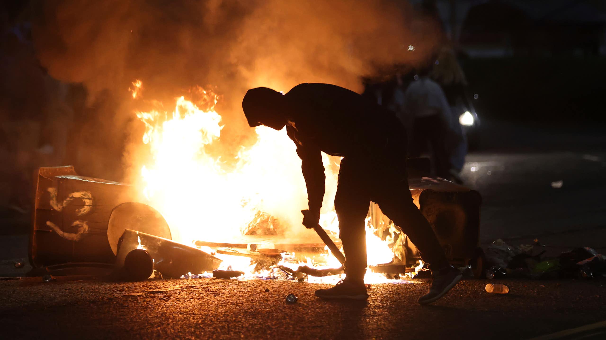 A hooded man bends as he stokes a fire.
