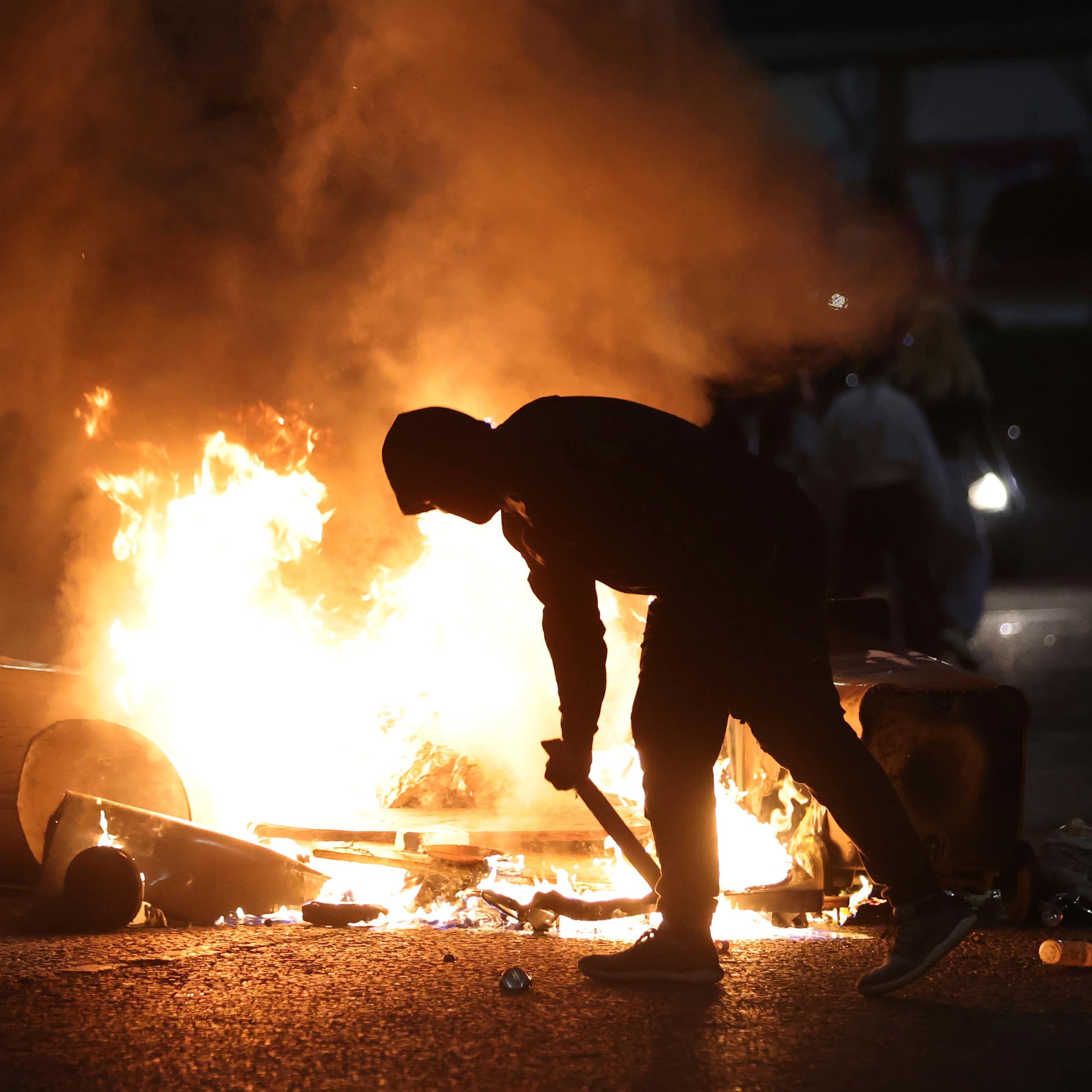 A hooded man bends as he stokes a fire.