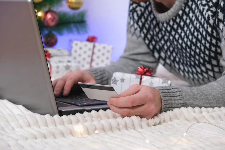 man in a christmas jumper with a christmas tree in the background making a purchase on his laptop with a credit card.