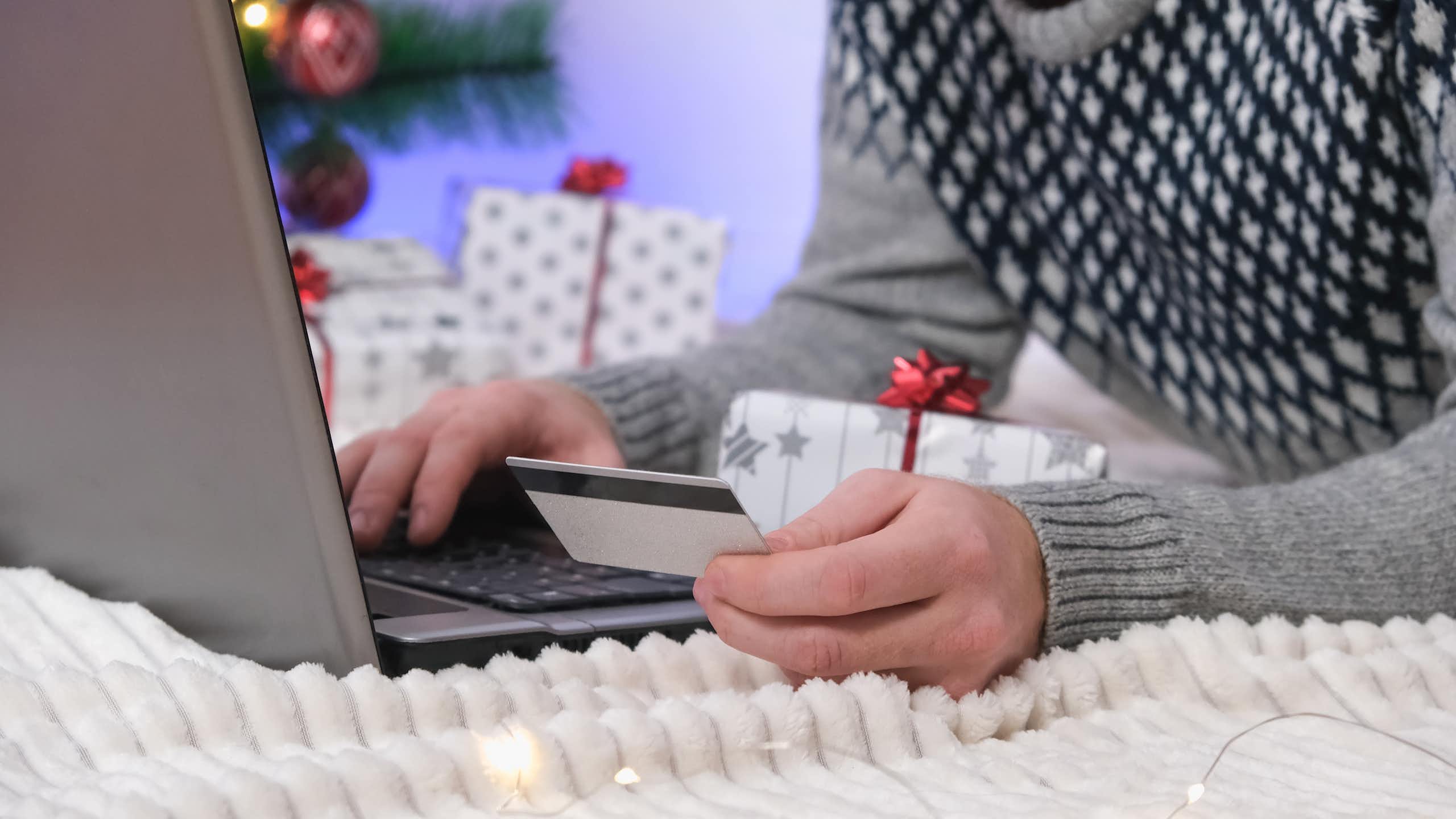 man in a christmas jumper with a christmas tree in the background making a purchase on his laptop with a credit card.