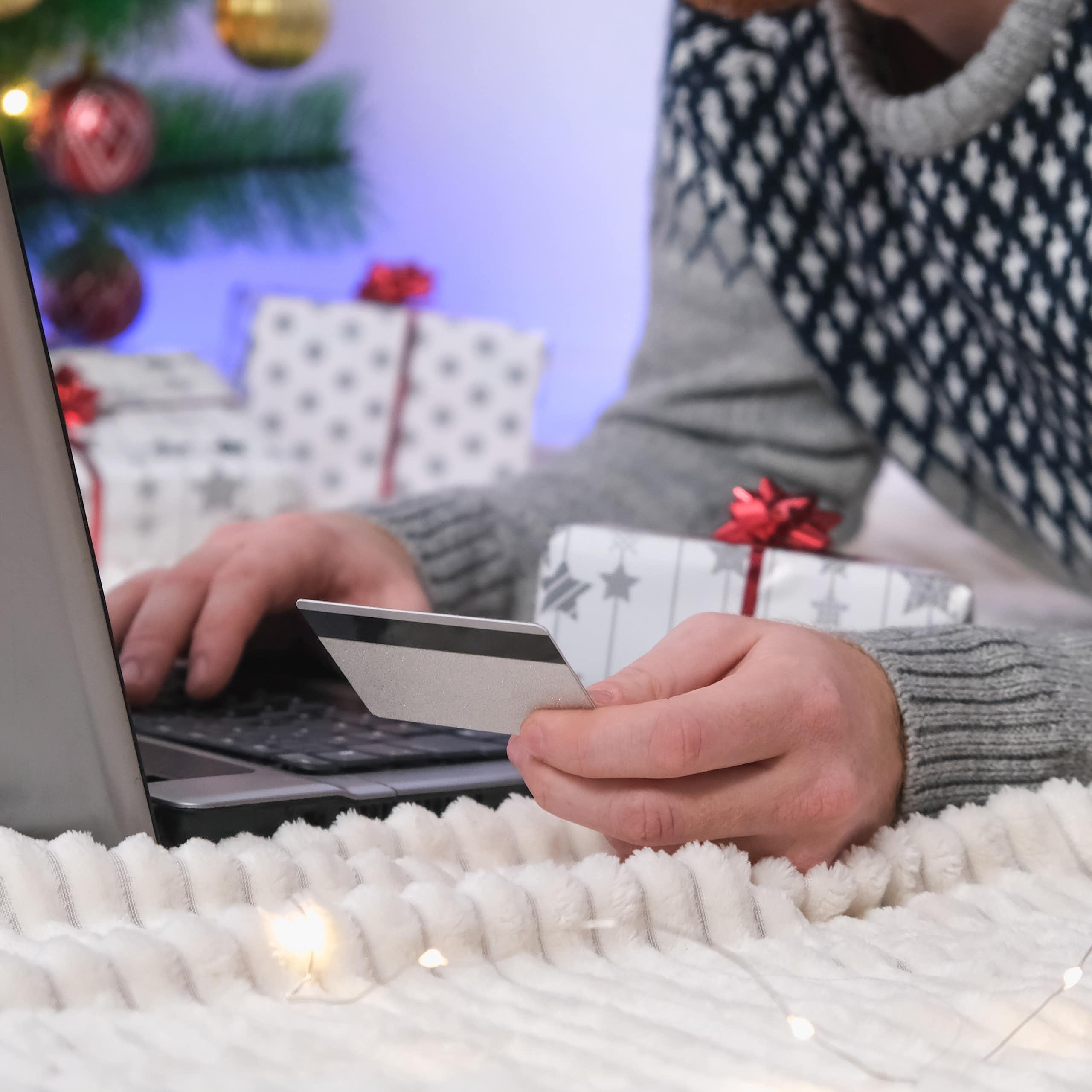 man in a christmas jumper with a christmas tree in the background making a purchase on his laptop with a credit card.