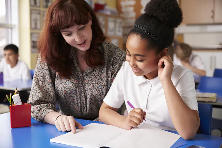 female teacher sitting beside a young girl at her desk, helping her with her work.