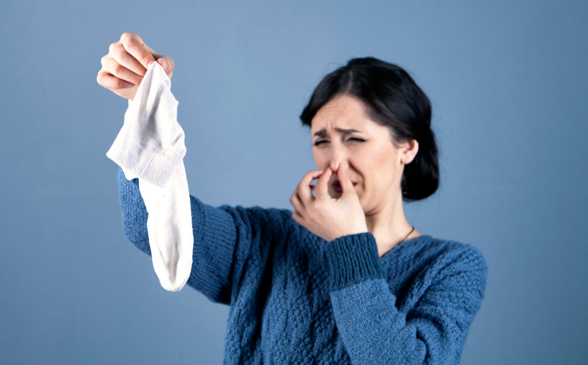 Uma mulher tapa o nariz e faz uma careta, olhando para uma meia branca malcheirosa que segura na outra mão.