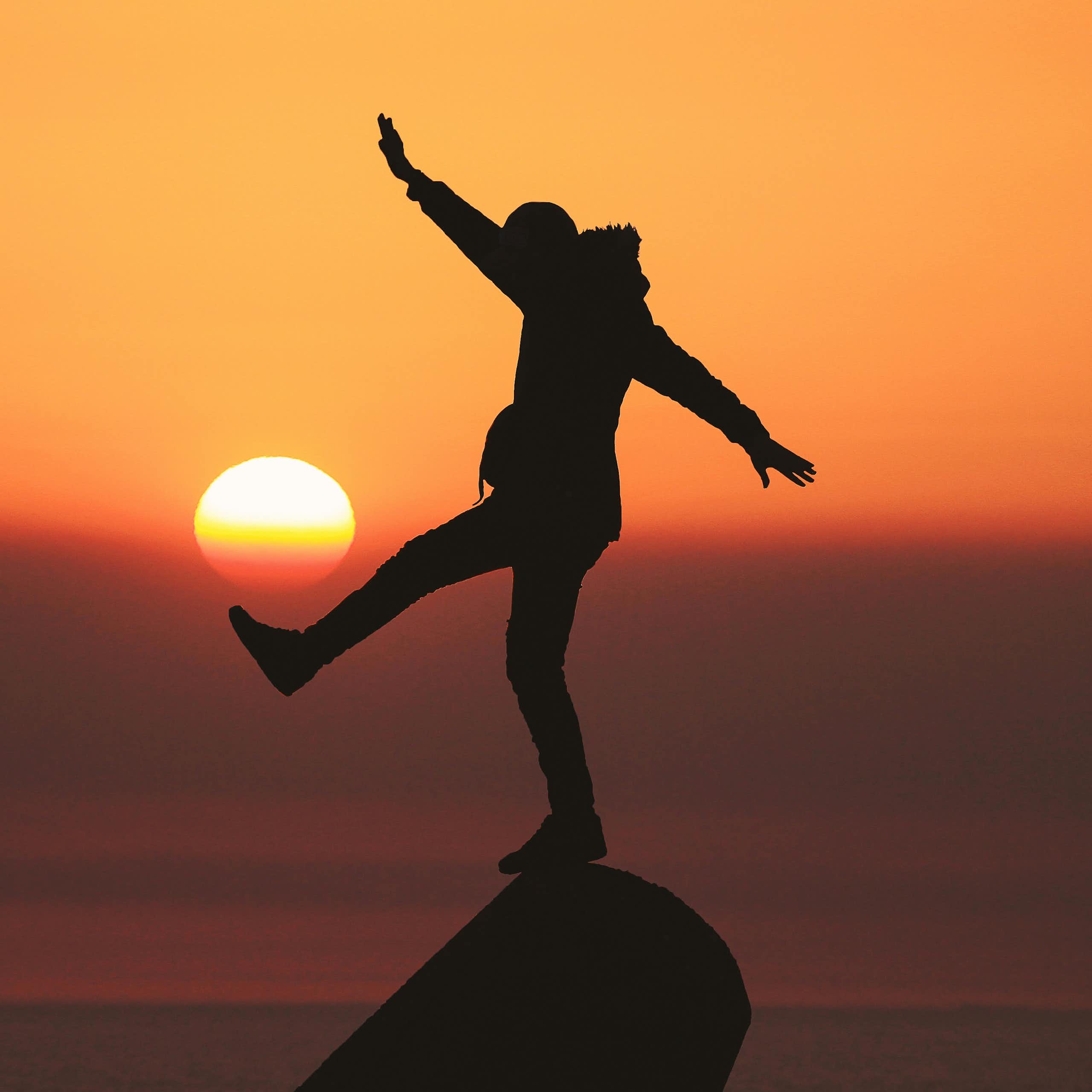 A person balances on a rock as the sun sets behind them.