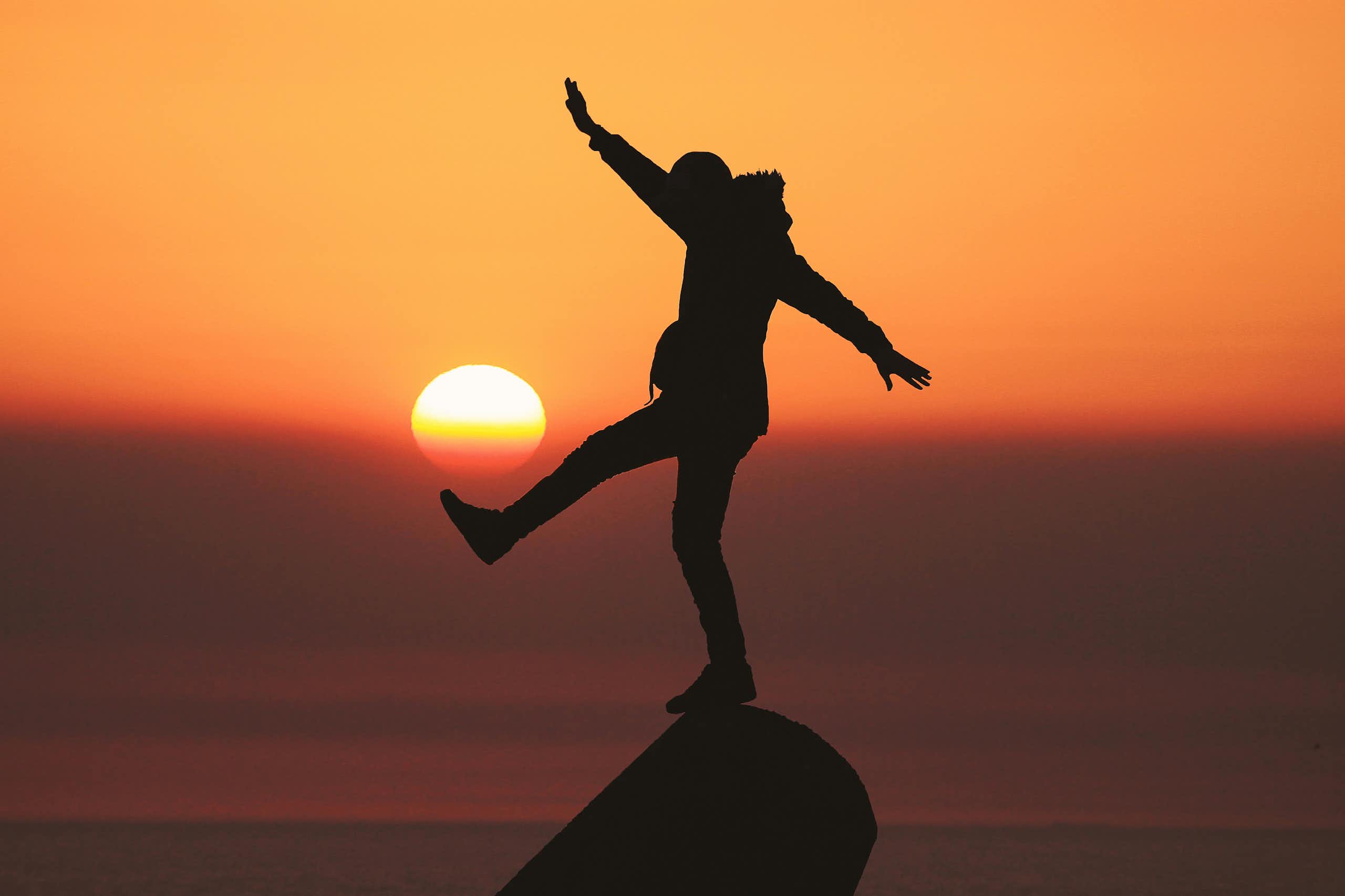 A person balances on a rock as the sun sets behind them.