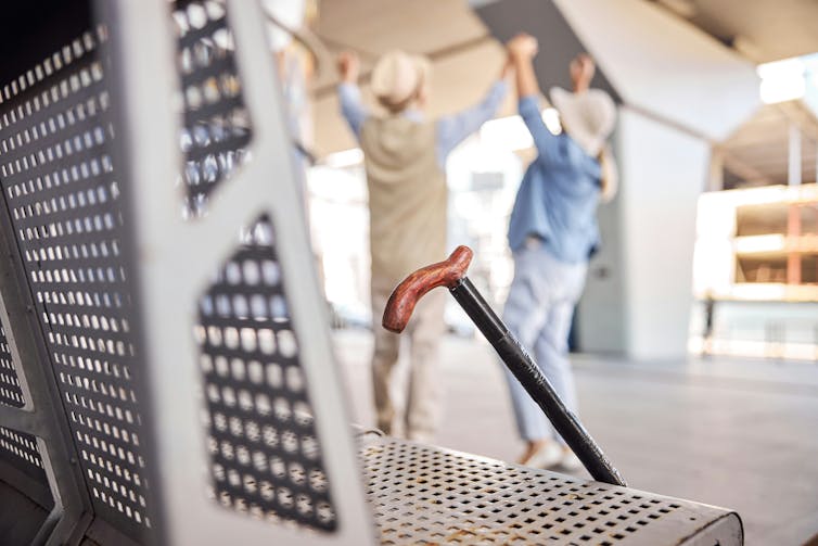 A cane leans against a bench as two older women stand with their arms raised a few feet away.