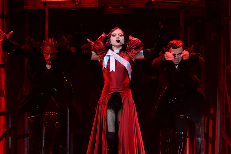 A performer (Lady Gaga) stands centre-stage in a dramatic red gown with black gloves, arms raised. Two dancers stand behind her, mid-movement. The stage is lit with deep red tones.