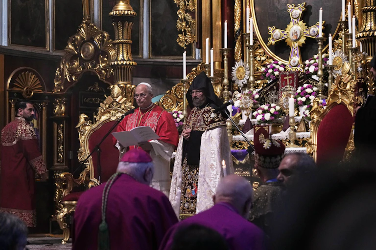 A man in an intricate robe stands beside another man while reading from a text. In the background, an altar with a crucifix and candles is visible.
