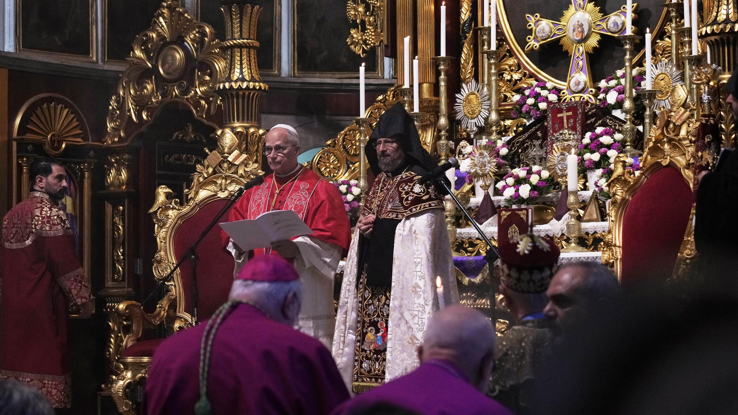 A man in an intricate robe stands beside another man while reading from a text. In the background, an altar with a crucifix and candles is visible.