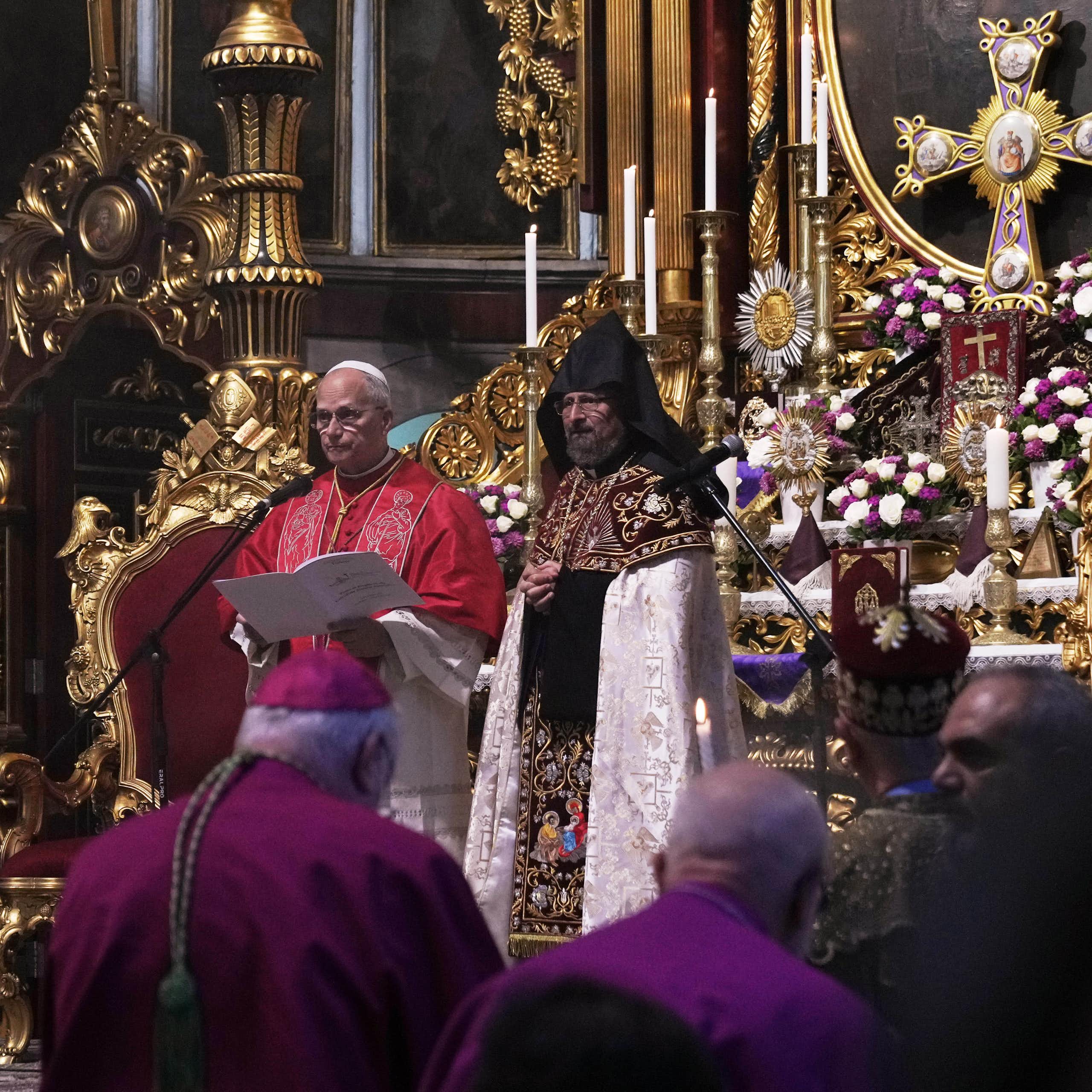 A man in an intricate robe stands beside another man while reading from a text. In the background, an altar with a crucifix and candles is visible.
