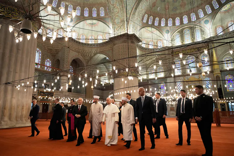 Several people walking in a large dome-like structure with high ceilings. Some are dressed in black suits and a few in white robes.