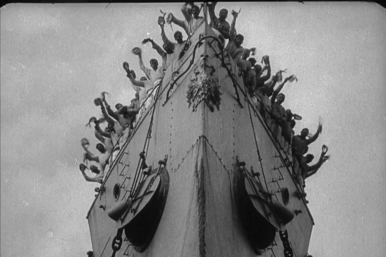 Black and white film still. Men wave over the bow of a ship.
