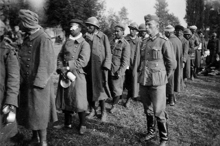 A black and white photo of African men in trenchcoats standing in a line with a European man in the foreground.