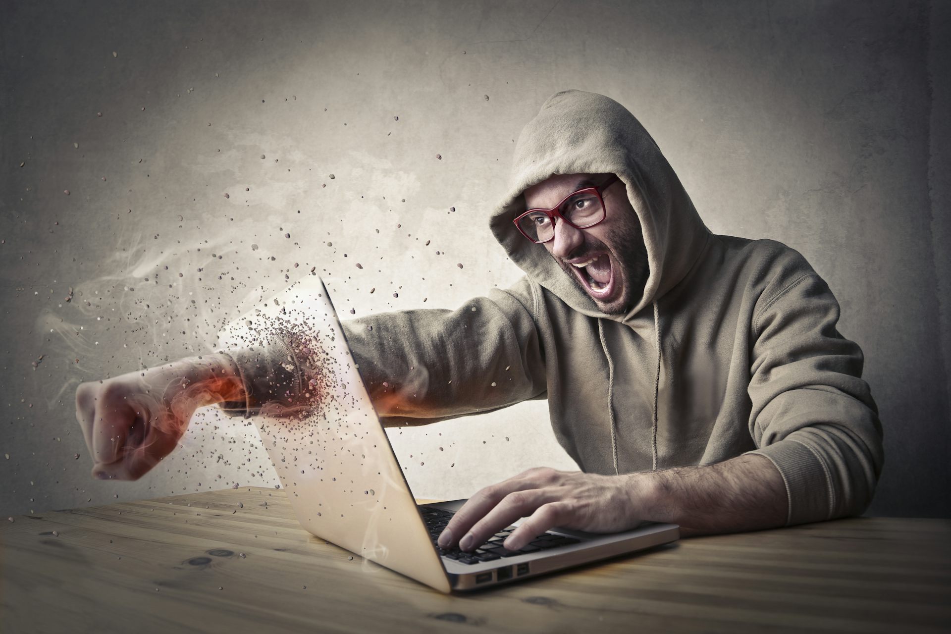 Man in hoodie smashing through laptop screen with fist.