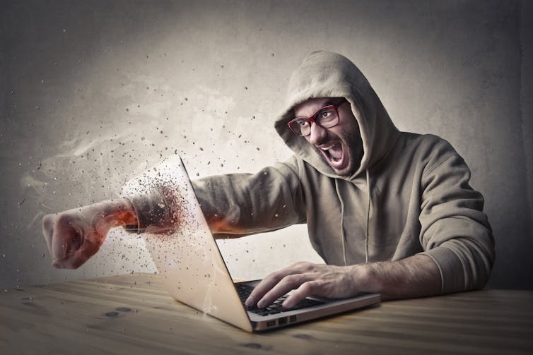 Man in hoodie smashing through laptop screen with fist.
