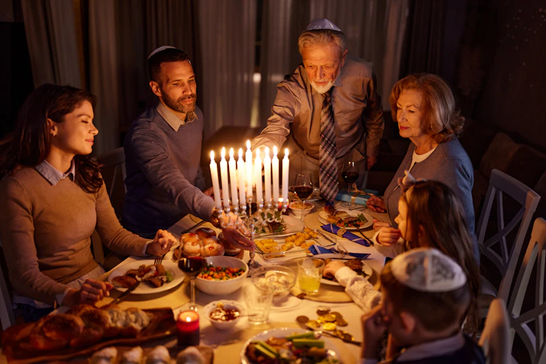 A family of four sits around the dinner table as an elderly man lights the menorah candles.