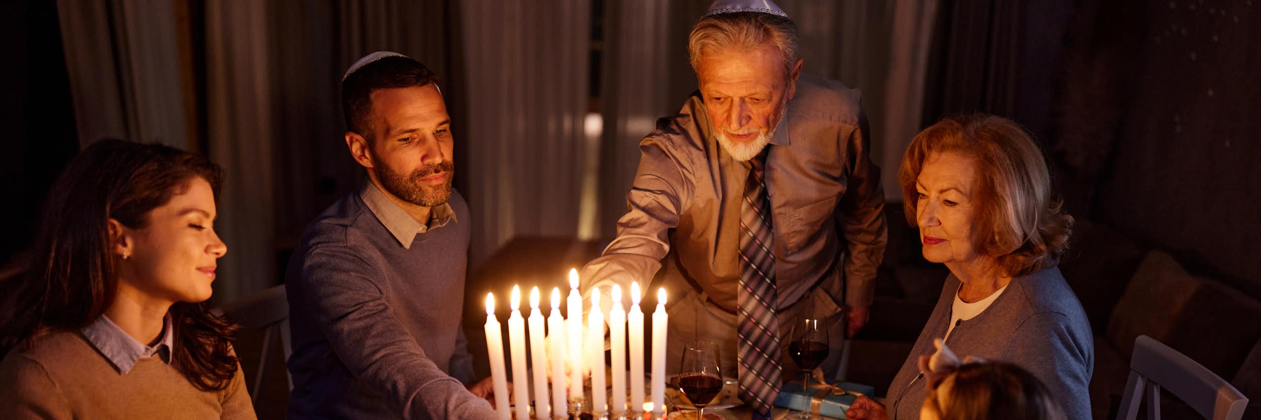 A family of four sits around the dinner table as an elderly man lights the menorah candles.