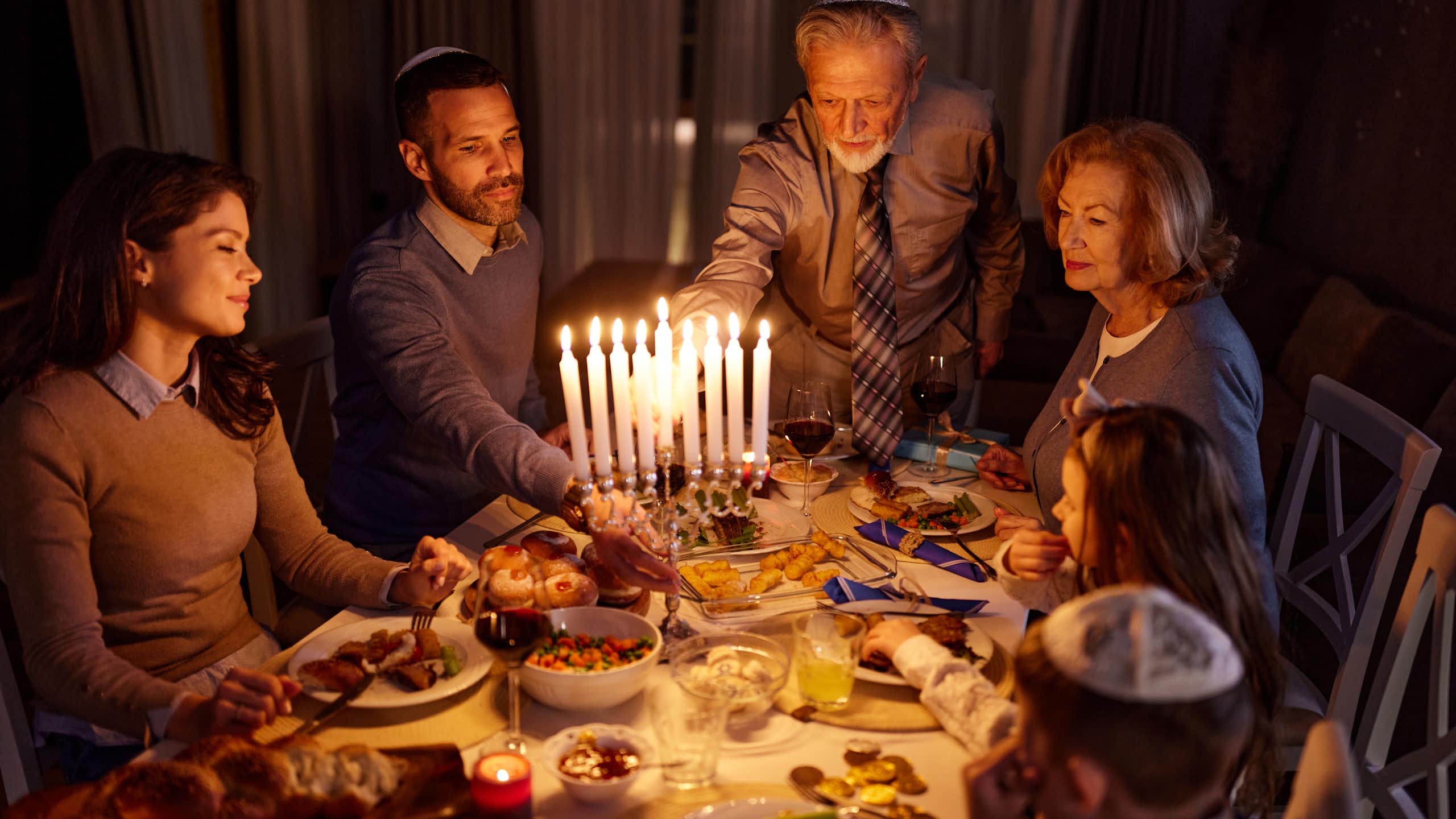 A family of four sits around the dinner table as an elderly man lights the menorah candles.