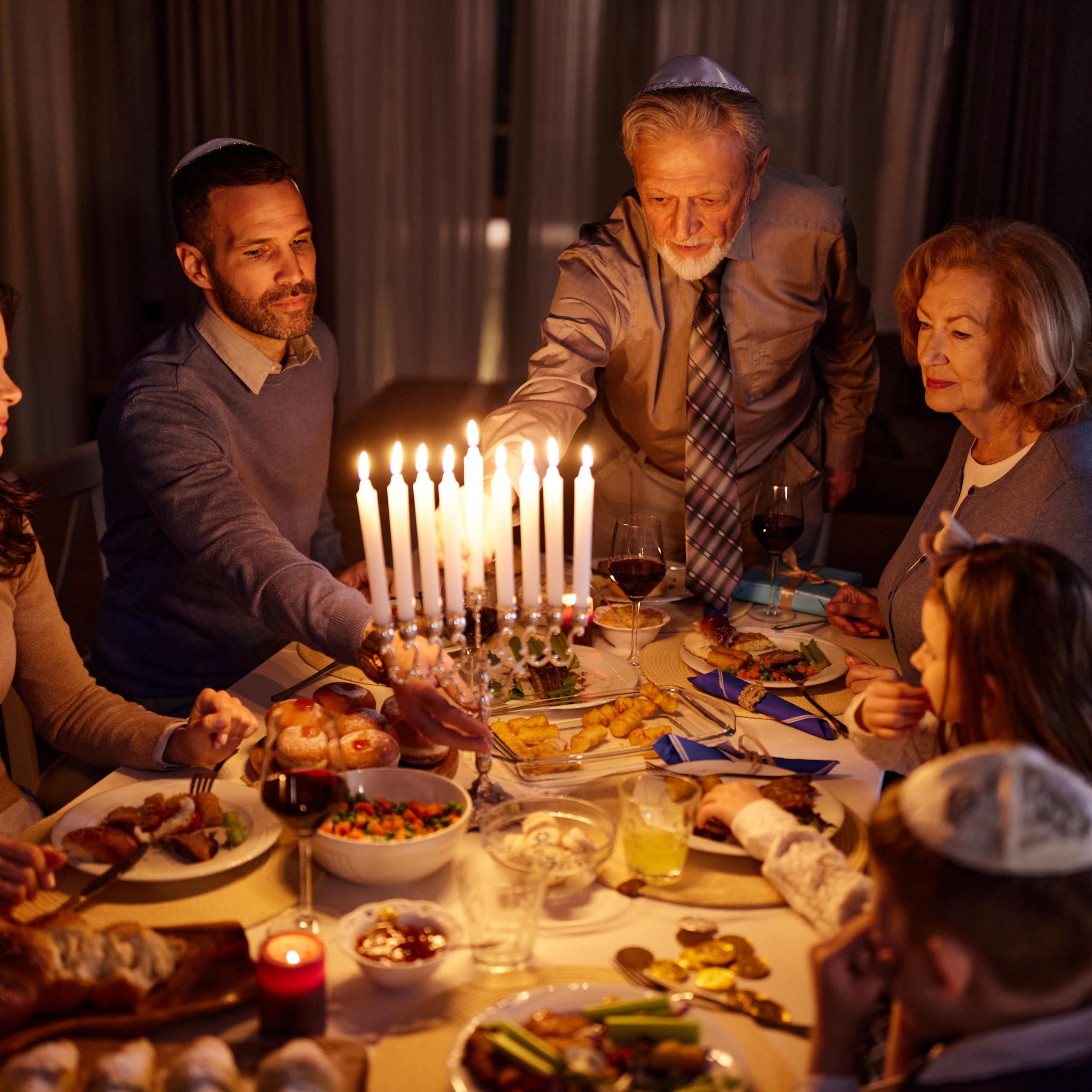 A family of four sits around the dinner table as an elderly man lights the menorah candles.