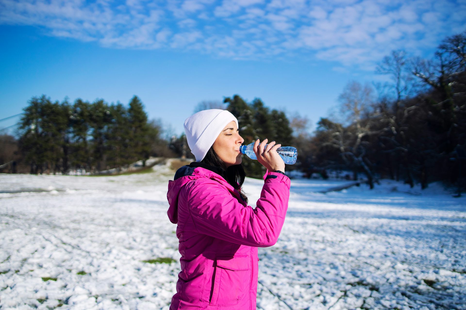 A woman wearing a white hat and a bright pink jacket sips from a water bottle while standing outside on a winter's day.